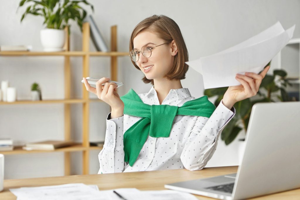 Portrait of talented female enterpreneur involved at working process, holds papers, makes voice call